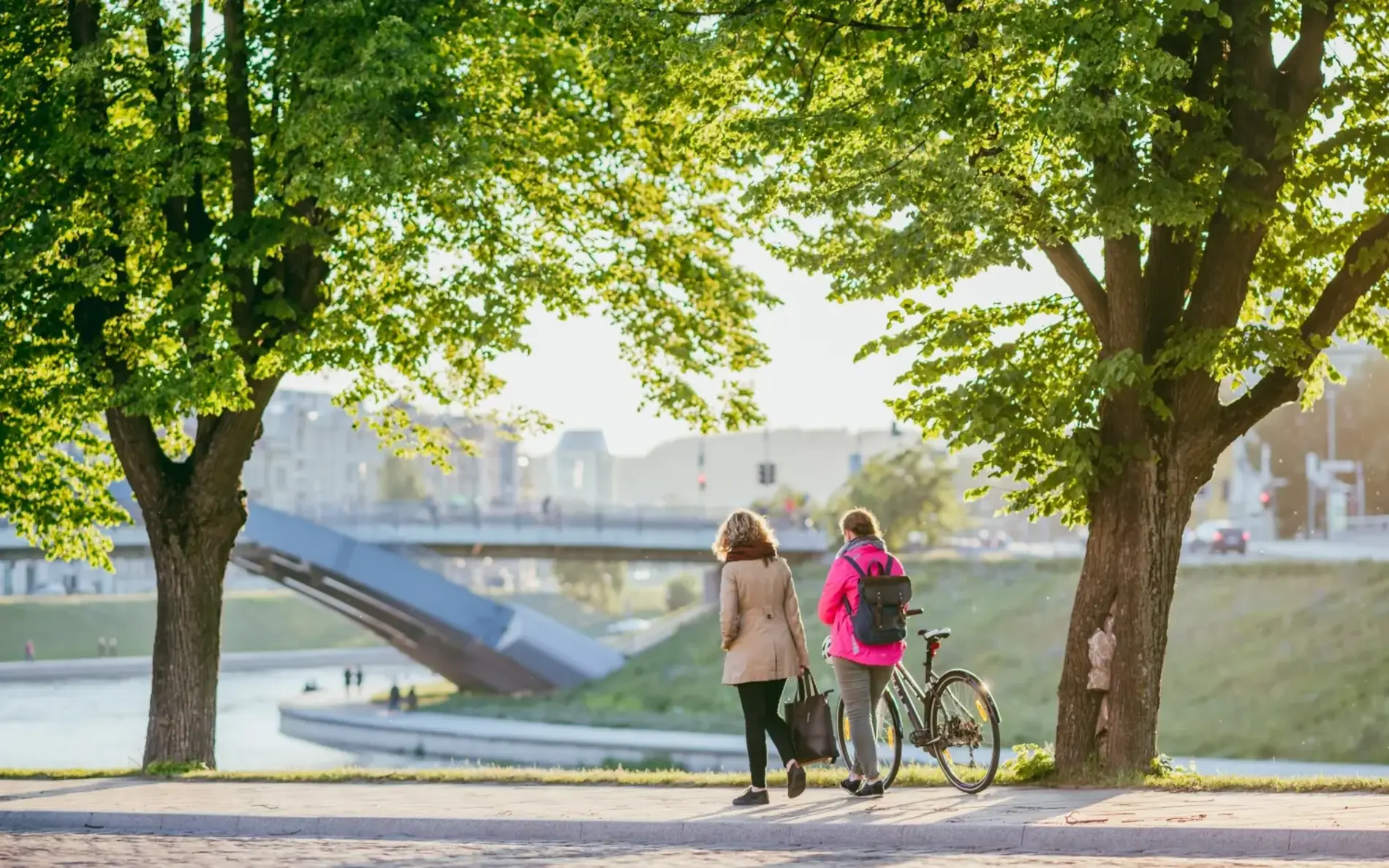 Zwei Frauen gehen auf einem Gehweg mit Bäumen, im Hintergrund eine große Metallbrücke. Eine Frau schiebt ihr Fahrrad, die andere geht daneben.