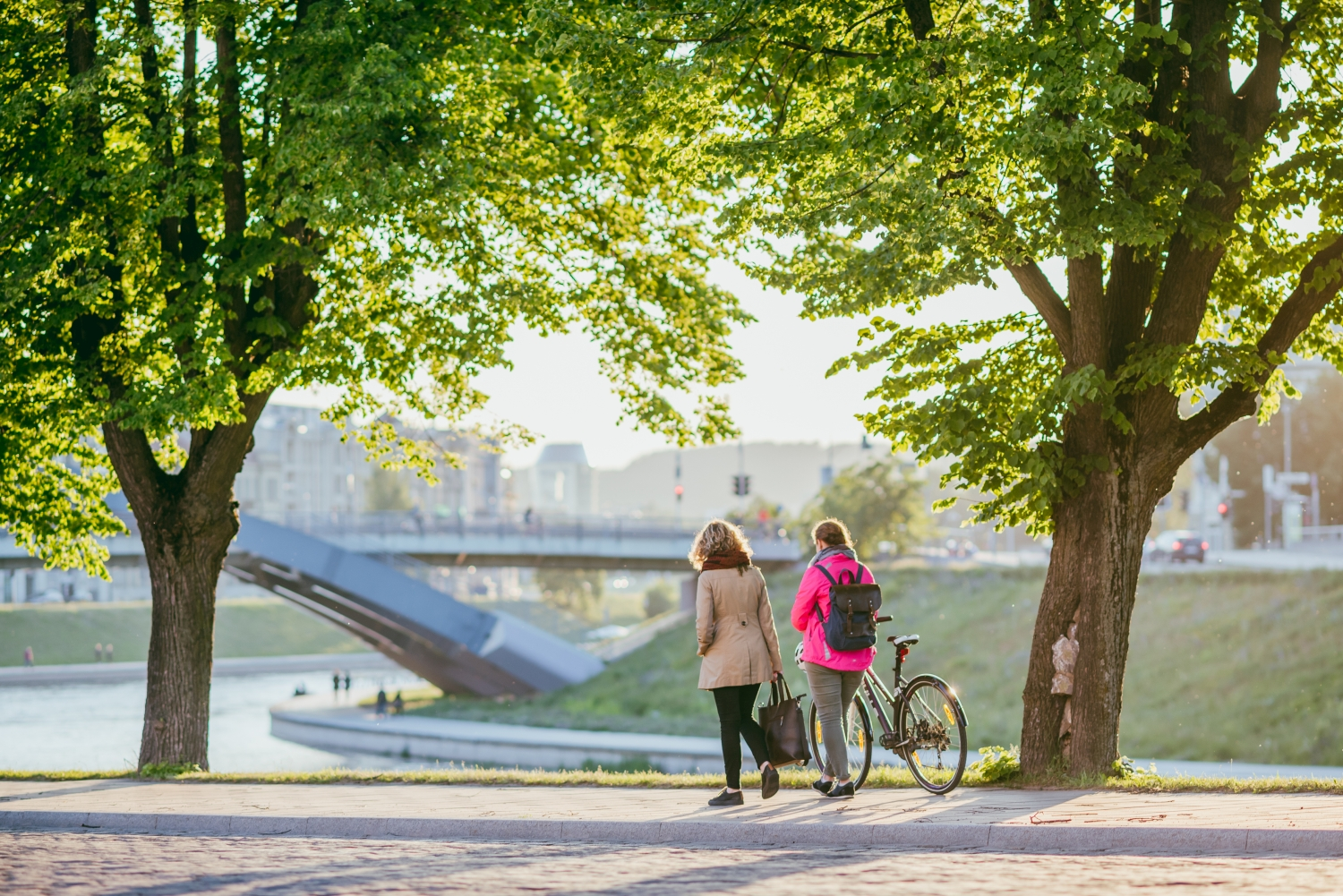 Zwei Frauen gehen auf einem Gehweg mit Bäumen, im Hintergrund eine große Metallbrücke. Eine Frau schiebt ihr Fahrrad, die andere geht daneben.