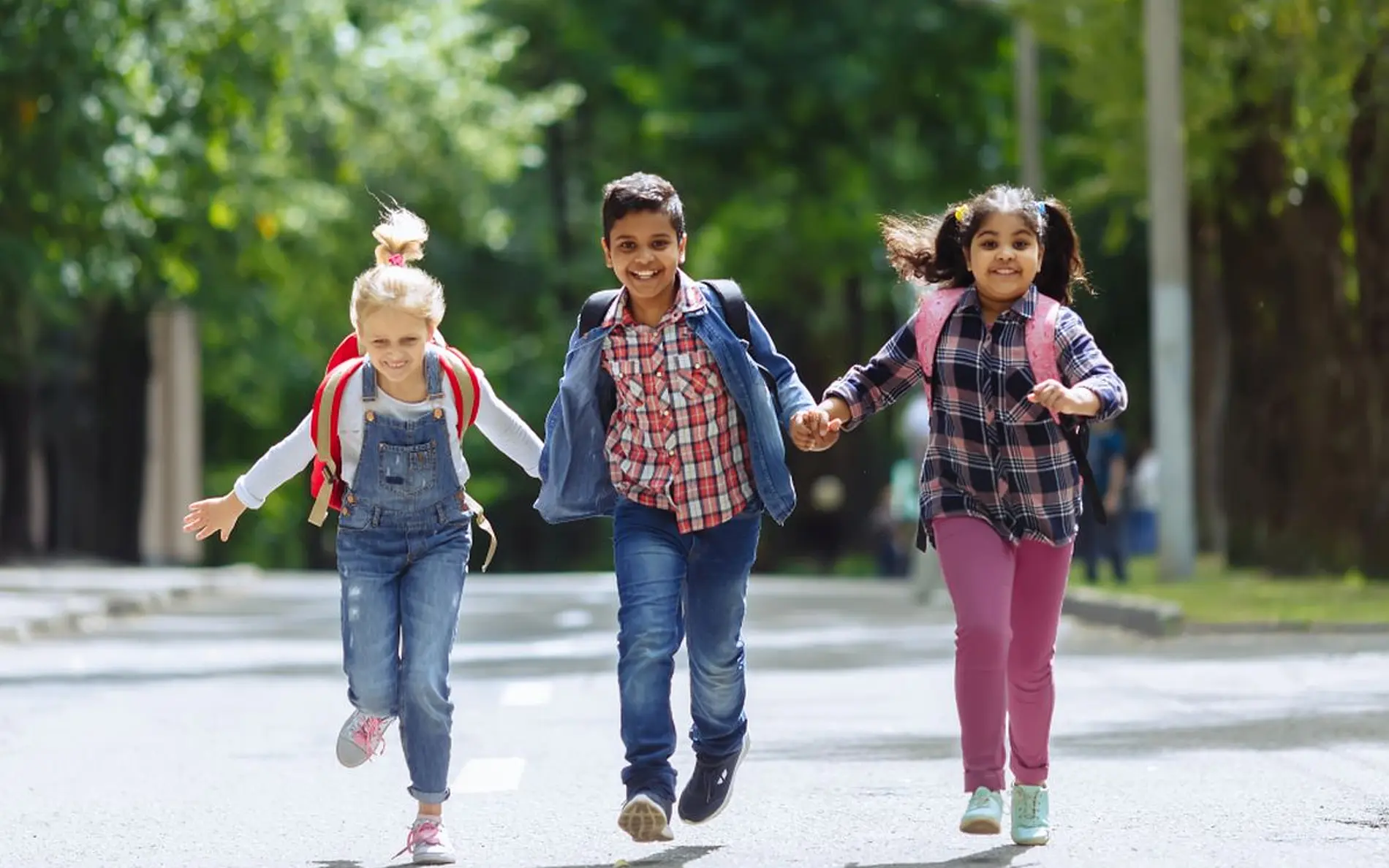 Gruppe von drei Kindern laufen Hand in Hand einen Weg entlang und lachen dabei. Sie tragen Schultaschen