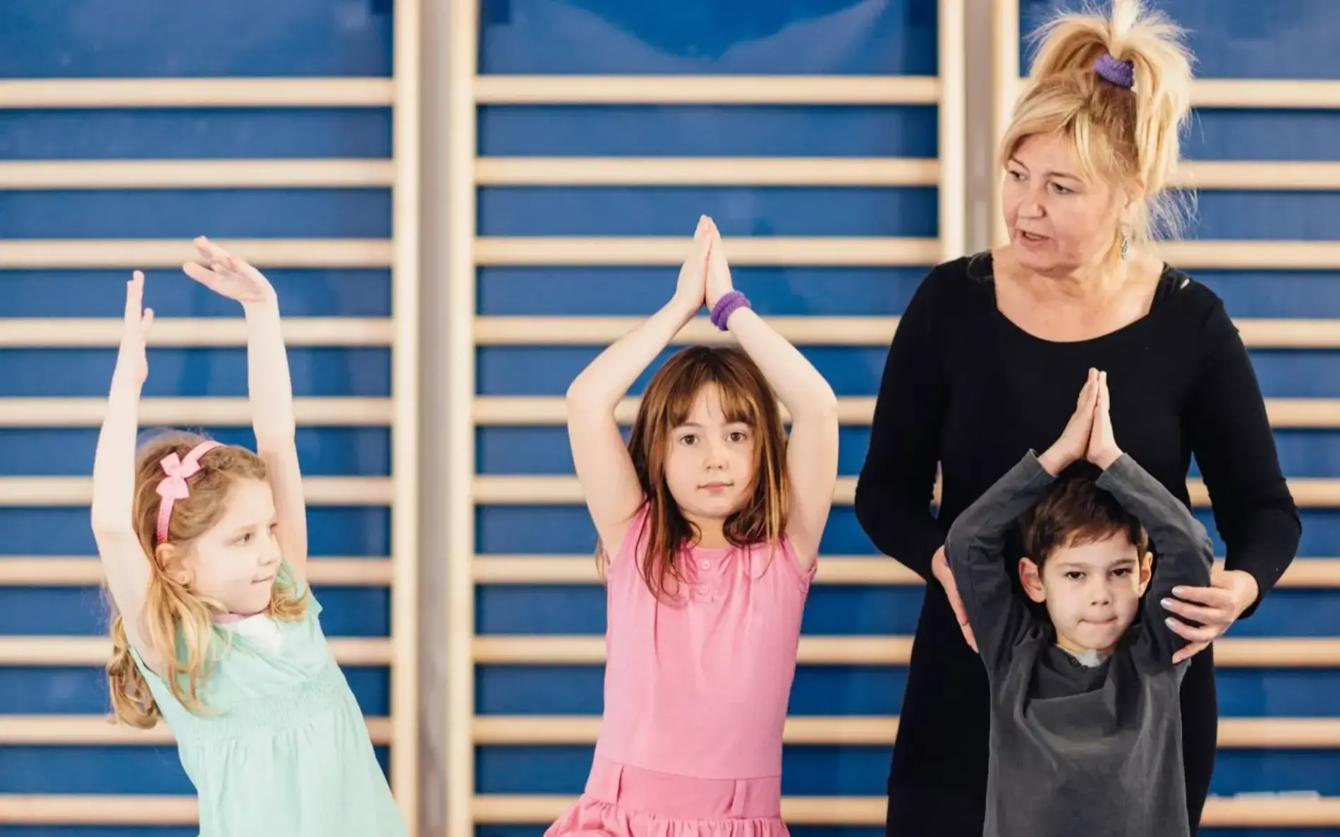 Drei Kinder und eine Lehrerin stehen vor einer Sprossenwand im Turnsaal. Die Kinder halten die Hände gefaltet über den Kopf und nehmen eine Yoga Pose ein