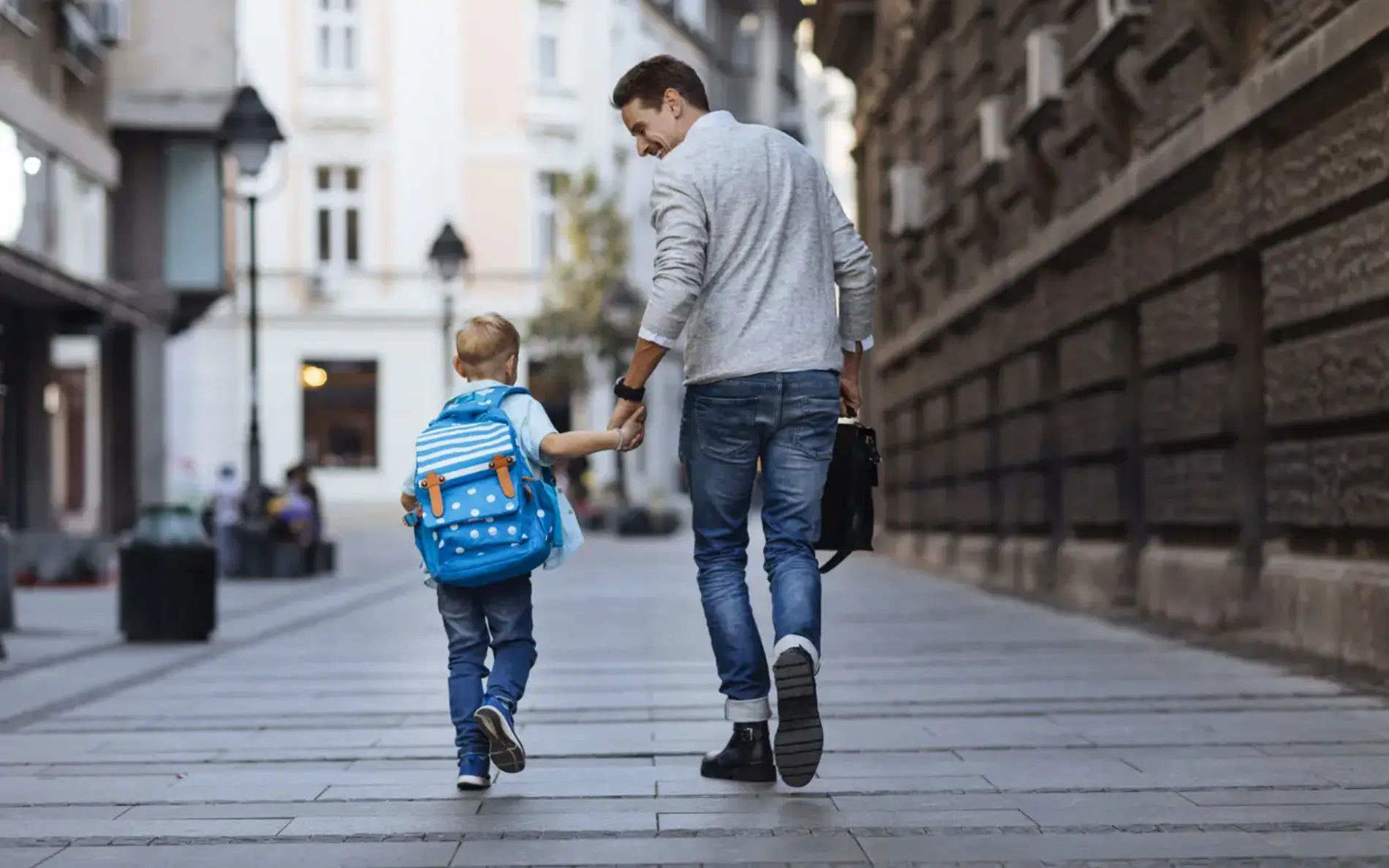 Ein Vater hält seinen Sohn mit Schultasche an der Hand. Sie gehen durch eine verkehrsberuhigte Straße in einer Gemeinde.
