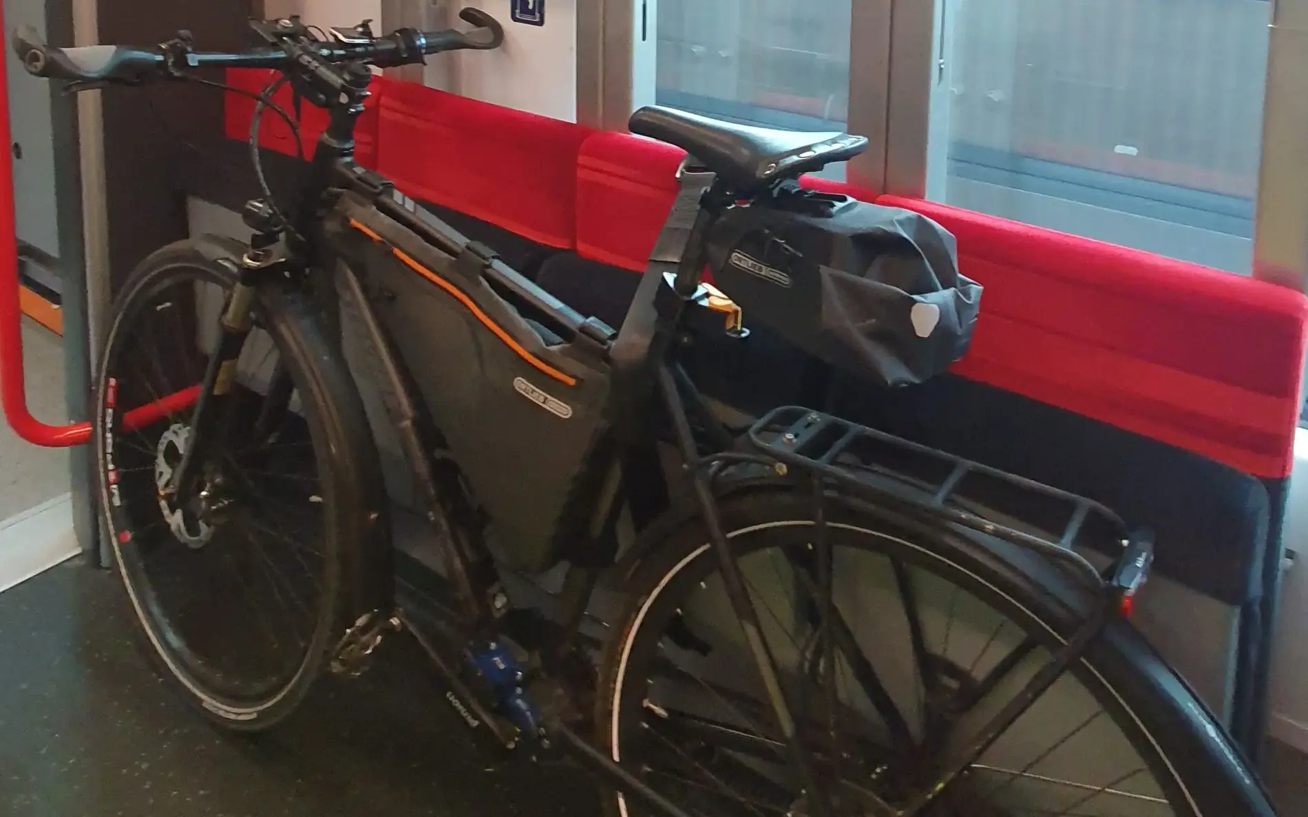 A black bicycle is secured inside a train in a designated bike area with red foldable seats and vertical metal bars. A sticker on the wall indicates priority space for bicycles and wheelchairs, and the train window shows blurred outdoor tracks.