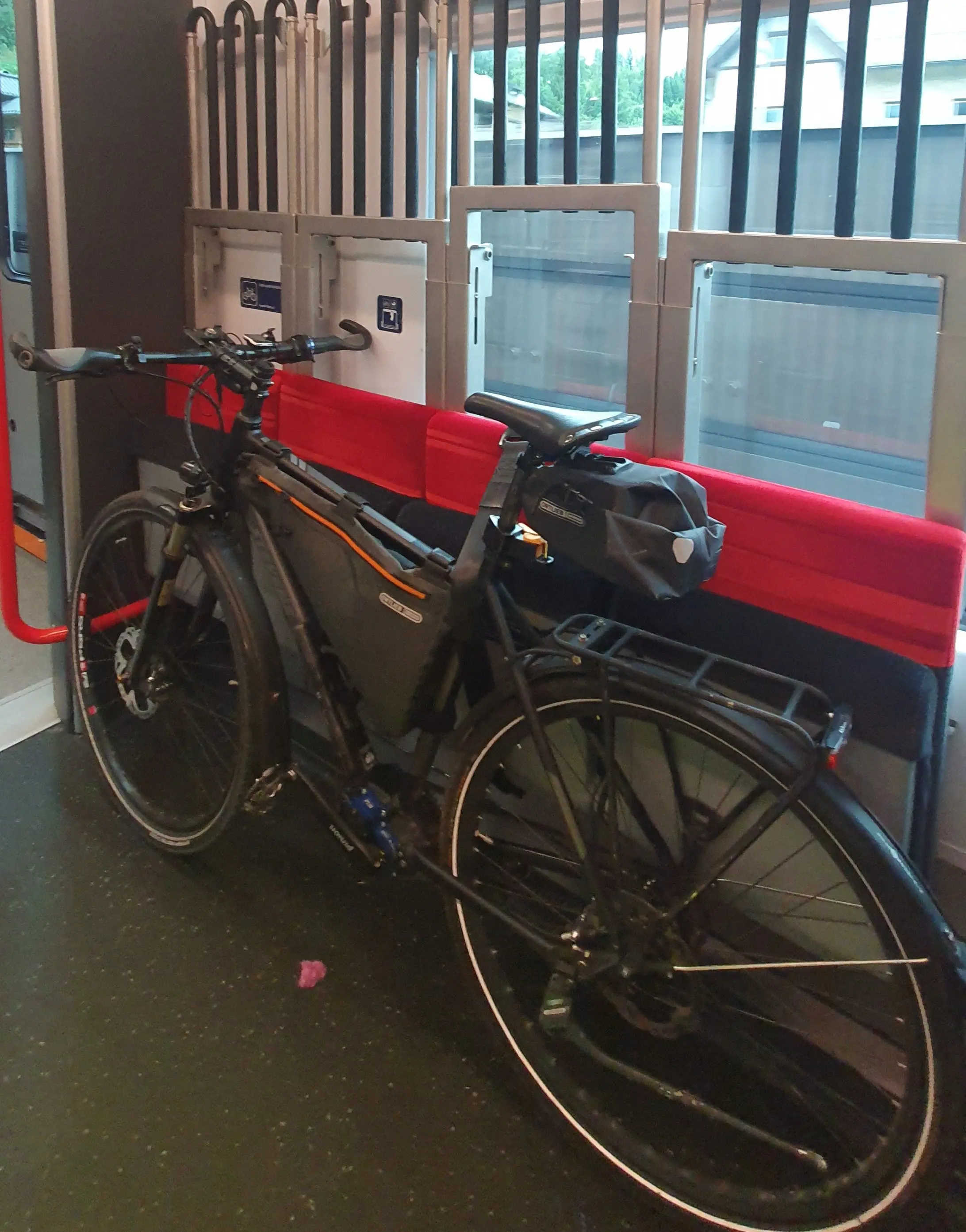 A black bicycle is secured inside a train in a designated bike area with red foldable seats and vertical metal bars. A sticker on the wall indicates priority space for bicycles and wheelchairs, and the train window shows blurred outdoor tracks.