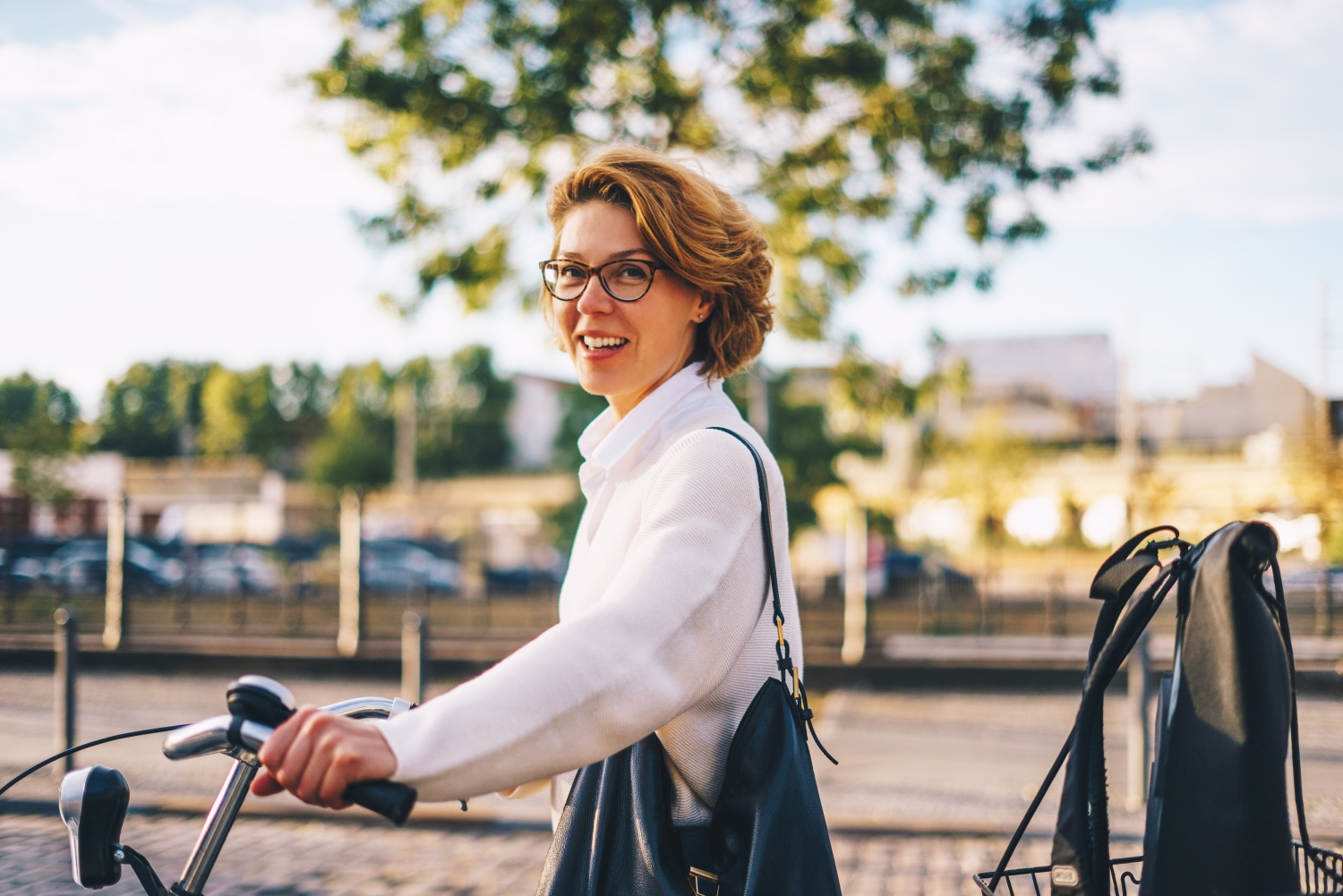 Eine Frau mit kurzen blonden Haaren, brille und weißem Hemd hat ihren Fahrradlenker in beiden Händen und lacht in die Kamera