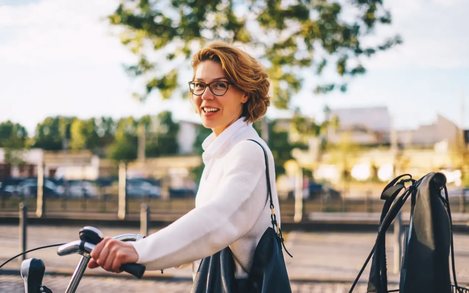 Eine Frau mit kurzen blonden Haaren, brille und weißem Hemd hat ihren Fahrradlenker in beiden Händen und lacht in die Kamera