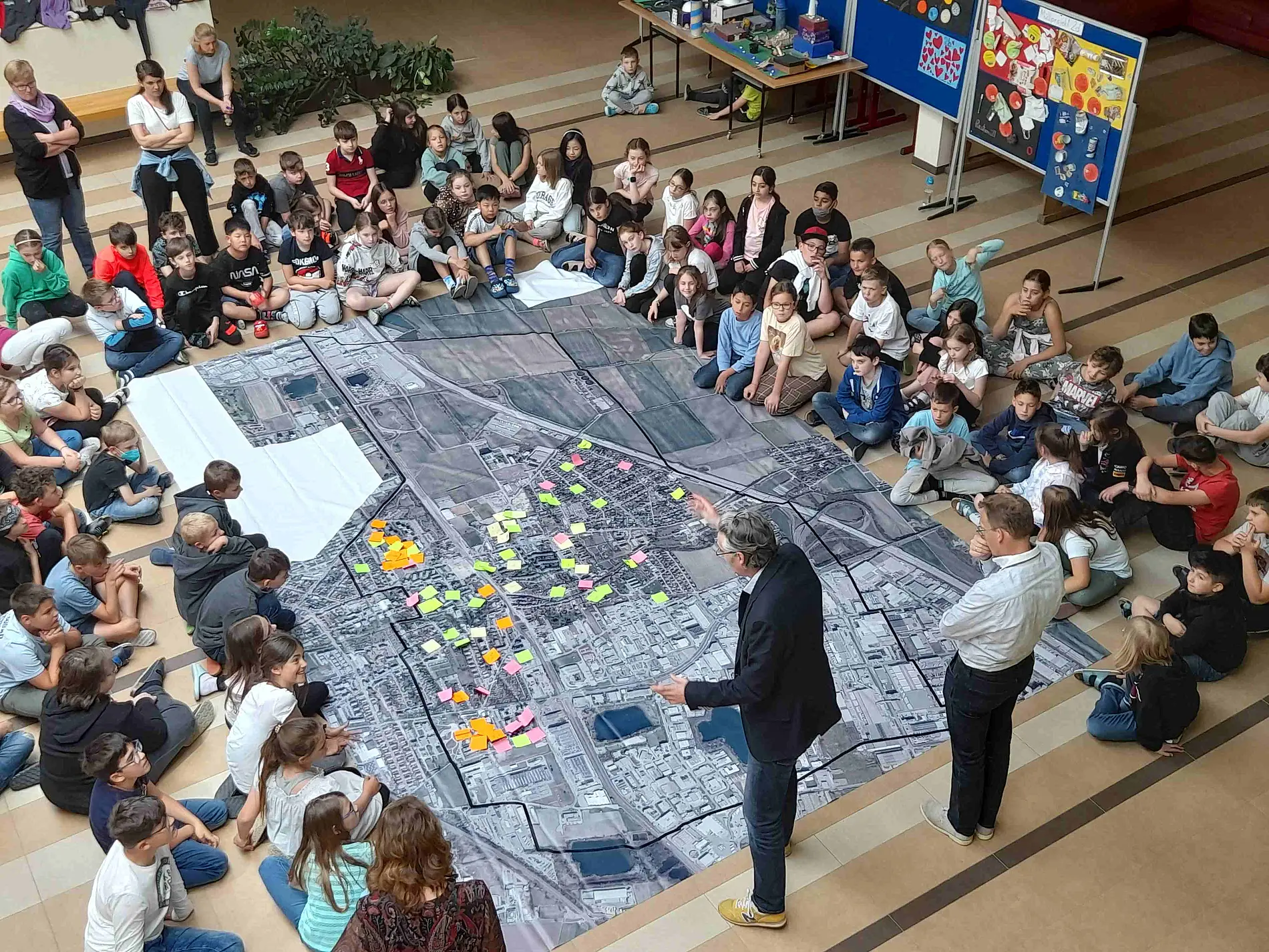 Große Gruppe Kinder sitzt am Boden rundum ein großes Luftbild der Gemeinde. Zwei Erwachsene stehen darauf und leiten an. Post-its kleben am Luftbild