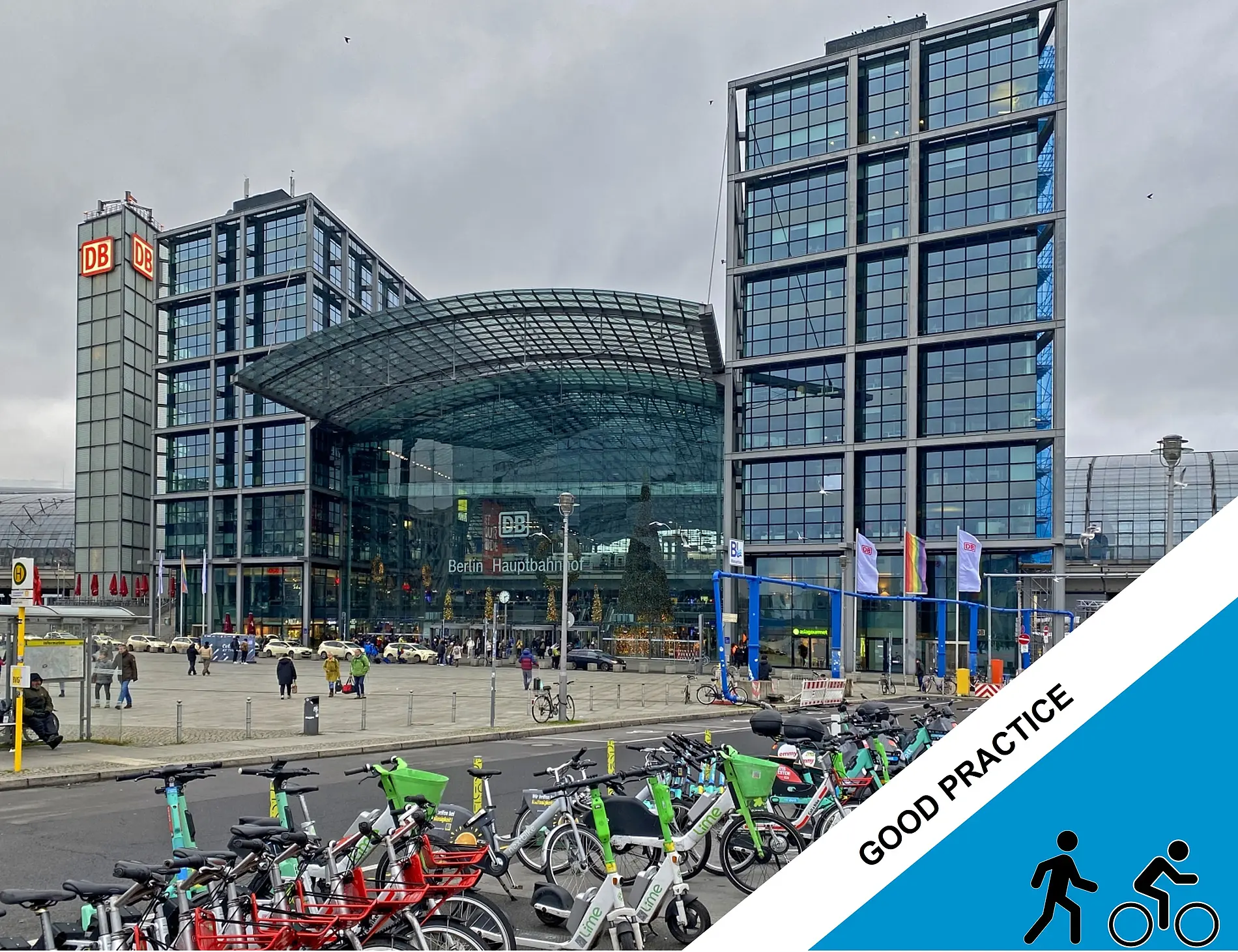 A large bicycle parking area with shared bicycles and e-scooters is situated in front of the glass-fronted Berlin Hauptbahnhof railway station, with the DB logo visible on the station buildings and people walking through the plaza.