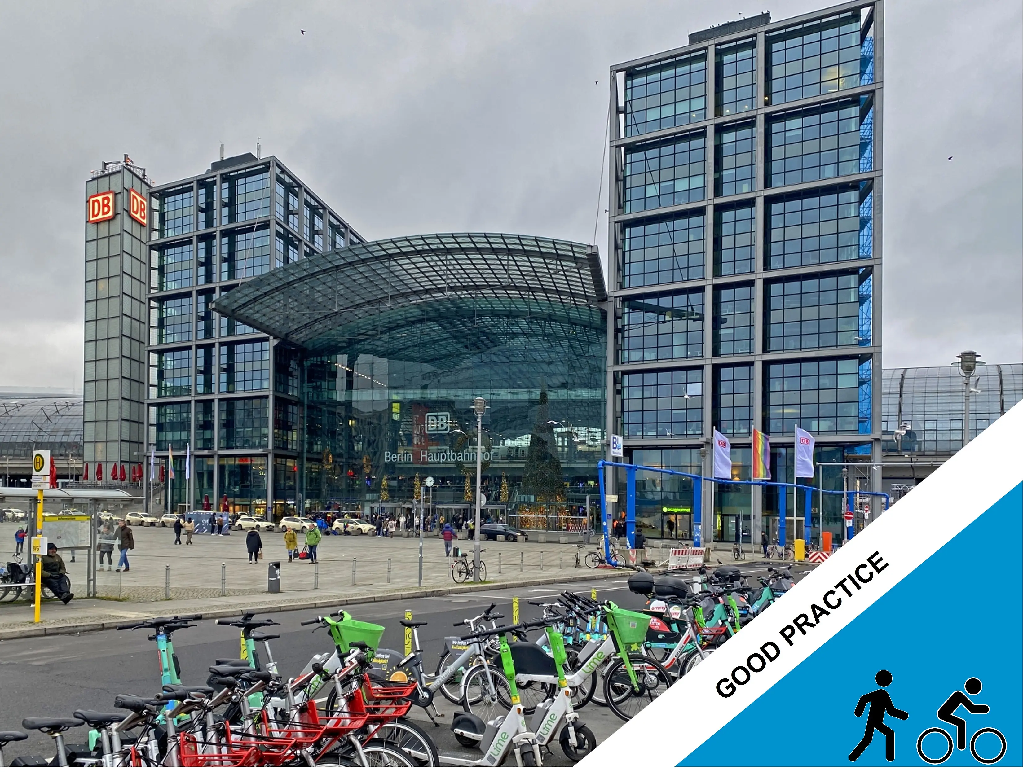 A large bicycle parking area with shared bicycles and e-scooters is situated in front of the glass-fronted Berlin Hauptbahnhof railway station, with the DB logo visible on the station buildings and people walking through the plaza.