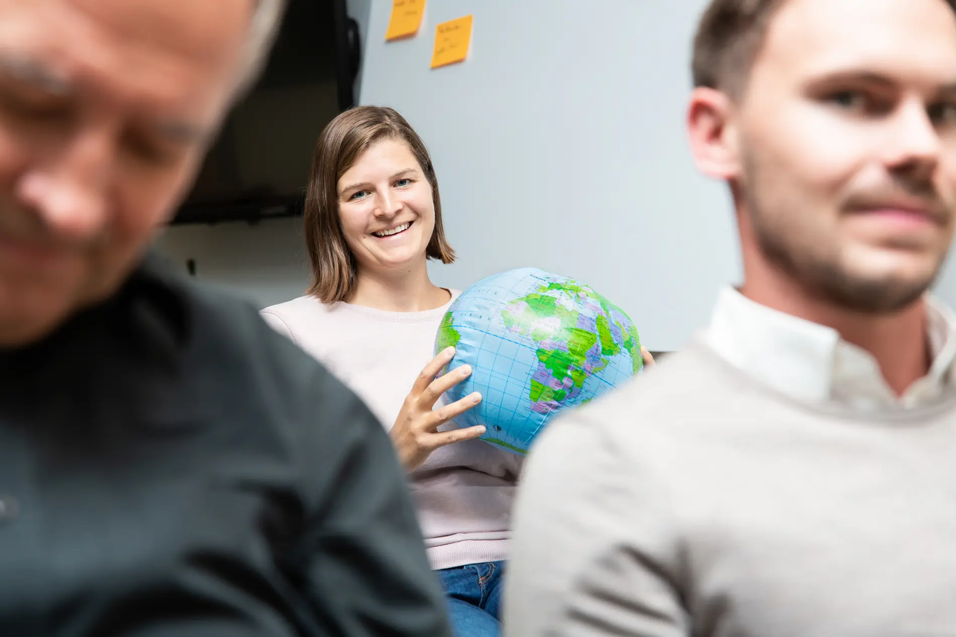 Anna Pribil hält einen Ball, der aussieht wie die Erde, und lacht in die Kamera. Sie sitzt hinter zwei Männern, die nicht im Fokus sind.
