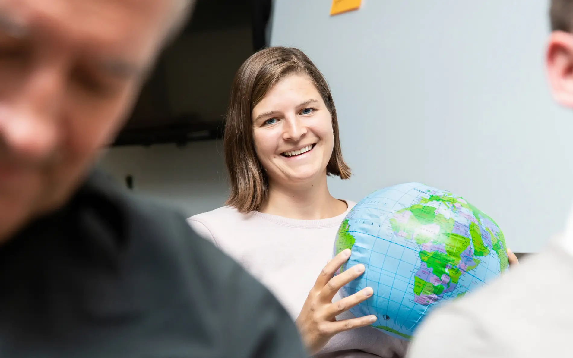 Anna Pribil hält einen Ball, der aussieht wie die Erde, und lacht in die Kamera. Sie sitzt hinter zwei Männern, die nicht im Fokus sind.