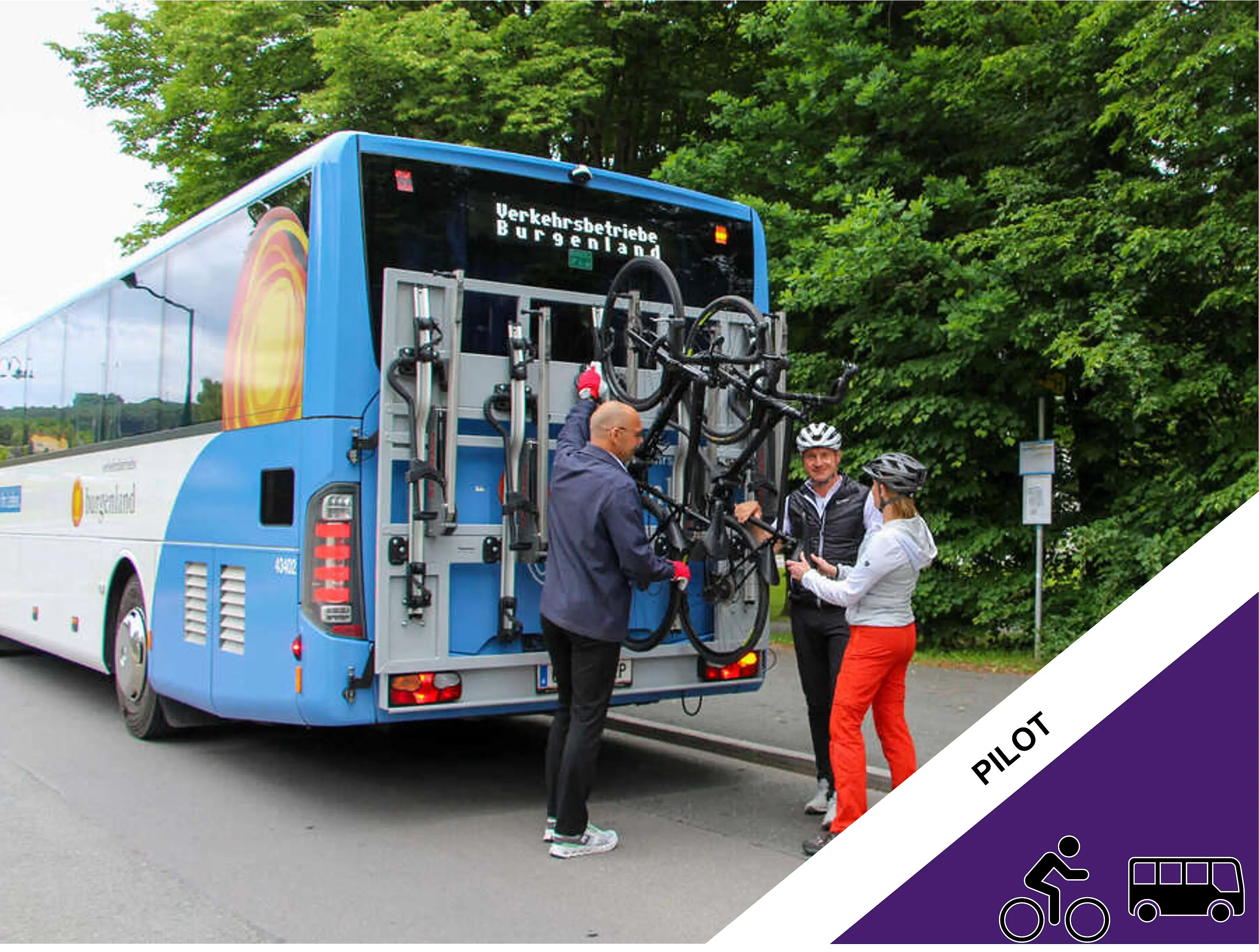 A blue bus is stopped at the roadside with a bicycle rack mounted on the back. Three people are attaching two bicycles to the rack. One person is lifting a bicycle into position, while two others guide and secure another bicycle. The bicycles are held upright and fastened at several points. In the background, there are green trees and the side of the bus with a large graphic design. The text “Verkehrsbetriebe Burgenland” is visible on the rear of the bus.