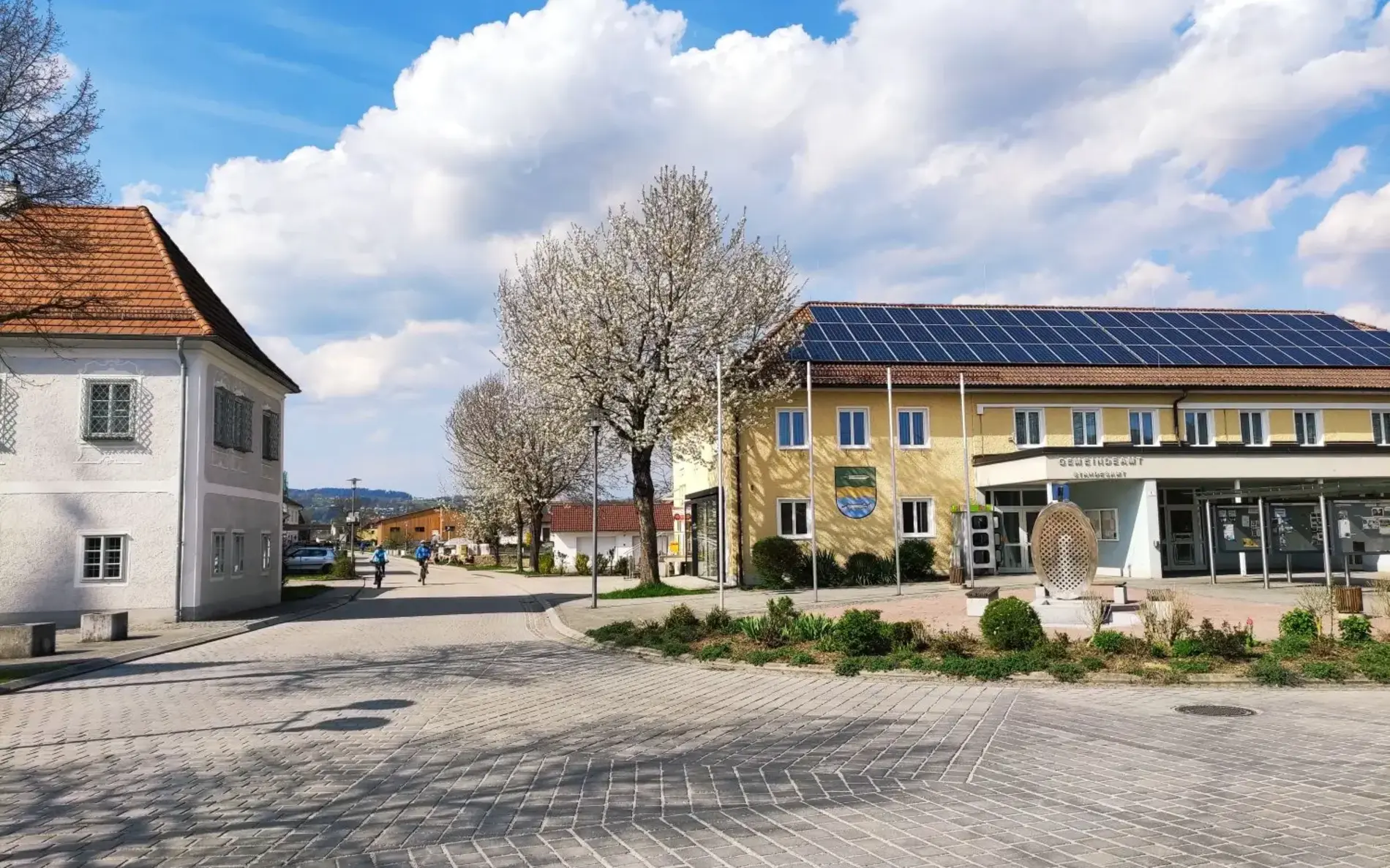 Das Foto zeigt ein Gemeindeamt mit Solarpanelen am Dach, davor ist dein Platz mit ein paar Bäumen und Begrünung.