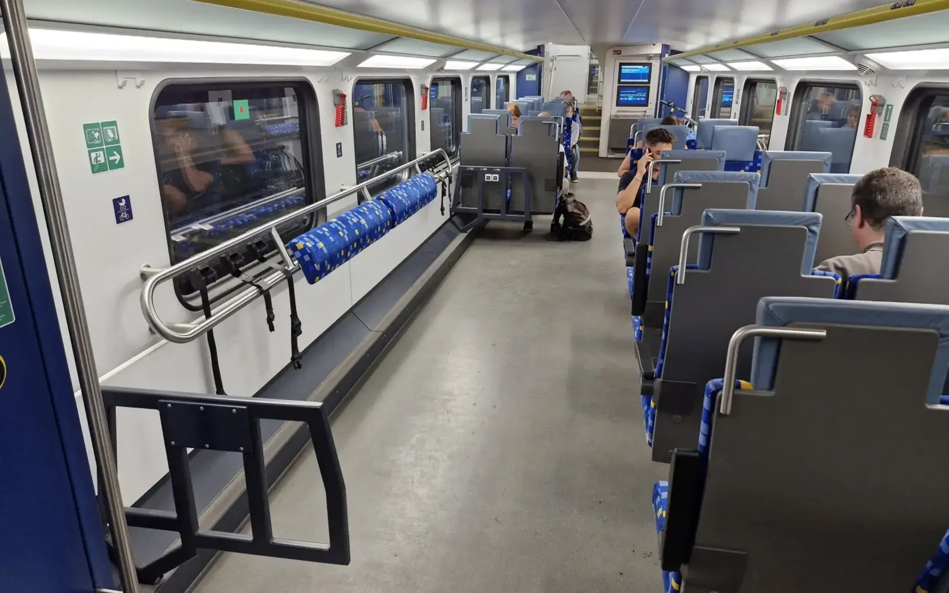 Das Foto zeigt das Innere eines modernen Doppelstockzuges mit breitem Mittelgang und Sitzreihen auf beiden Seiten. Links befindet sich ein Bereich für Fahrräder mit Halterungen und blauen Schutzpolstern. An den Wänden sind Notfall- und Sicherheitshinweise angebracht. Im Hintergrund ist ein Bildschirm mit Fahrgastinformationen sichtbar. Einige Personen sitzen auf den Sitzen, und auf dem Boden liegt eine Tasche