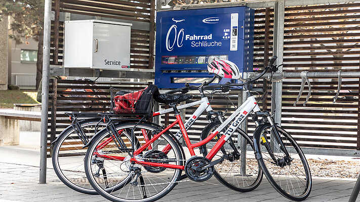 Zwei Räder mit dem Branding der TU Graz lehnen an einer überdachten Radservice-Box. Auf einem Automaten steht "Fahrrad Schläuche"