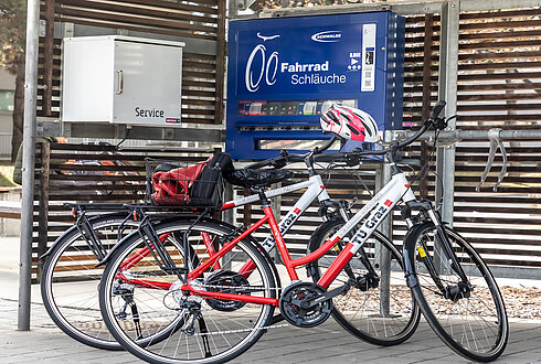 Zwei Räder mit dem Branding der TU Graz lehnen an einer überdachten Radservice-Box. Auf einem Automaten steht "Fahrrad Schläuche"
