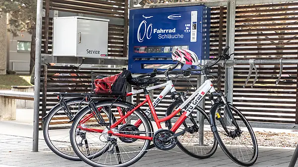 Zwei Räder mit dem Branding der TU Graz lehnen an einer überdachten Radservice-Box. Auf einem Automaten steht "Fahrrad Schläuche"