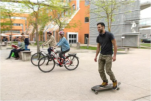 Ein Skateboardfahrer, zwei Radfahrende und im Hintergrund eine Person, die auf einer Bank sitzt.