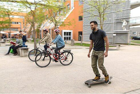 Ein Skateboardfahrer, zwei Radfahrende und im Hintergrund eine Person, die auf einer Bank sitzt.