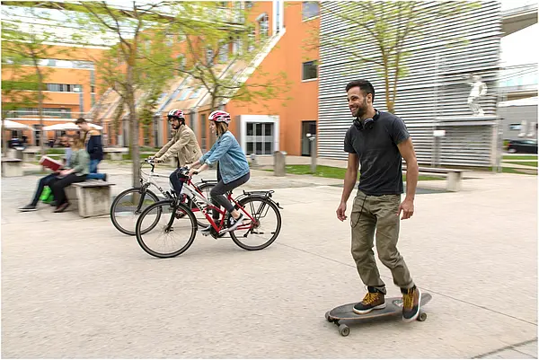 Ein Skateboardfahrer, zwei Radfahrende und im Hintergrund eine Person, die auf einer Bank sitzt.