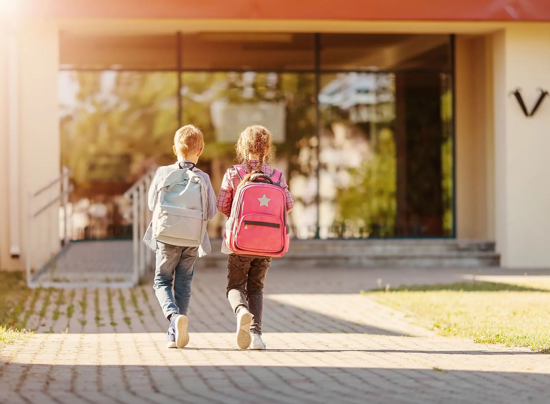 Zwei Kinder von hinten mit großen Schultaschen gehen einen gepflasterten Weg entlang, wo aus den Ritzen Grün ragt. Sie gehen auf ein Gebäude im Hintergrund zu 