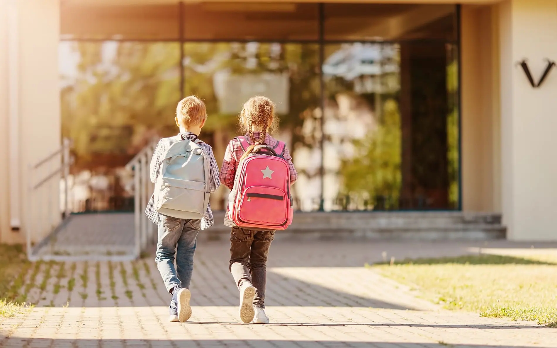 Zwei Kinder von hinten mit großen Schultaschen gehen einen gepflasterten Weg entlang, wo aus den Ritzen Grün ragt. Sie gehen auf ein Gebäude im Hintergrund zu 