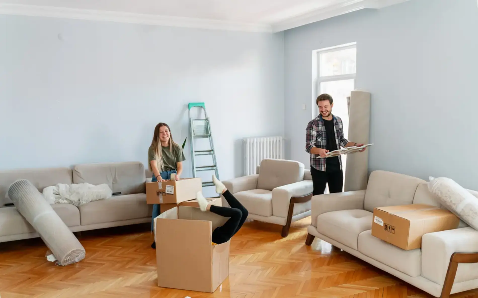 Eine Frau und ein Mann packen ihre Umzugskartons aus. Eine dritte Frau steckt kopfüber in einem Umzugskarton