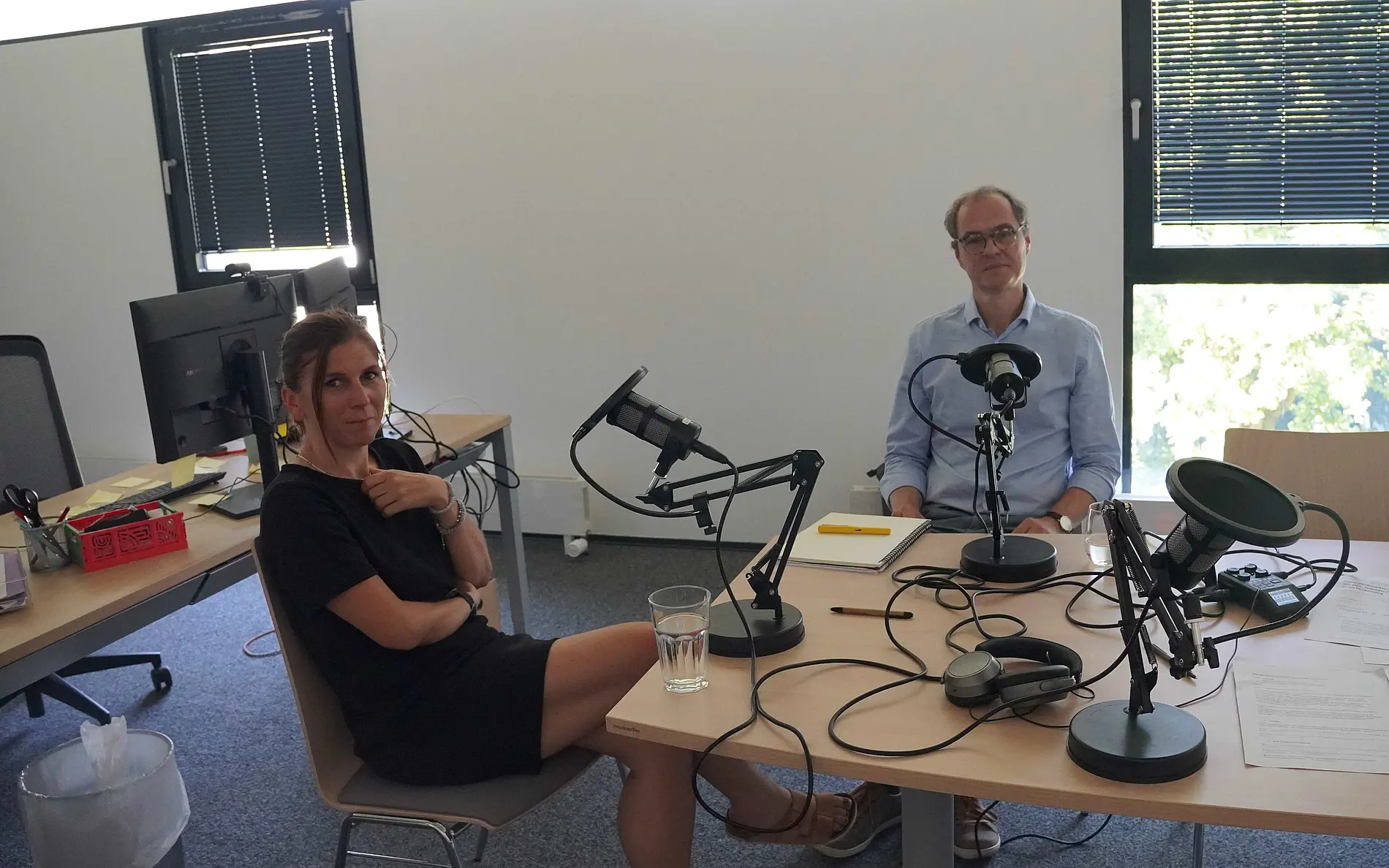 ein Mann und eine Frau vor Podcast-Mikrofonen lächeln in die Kamera