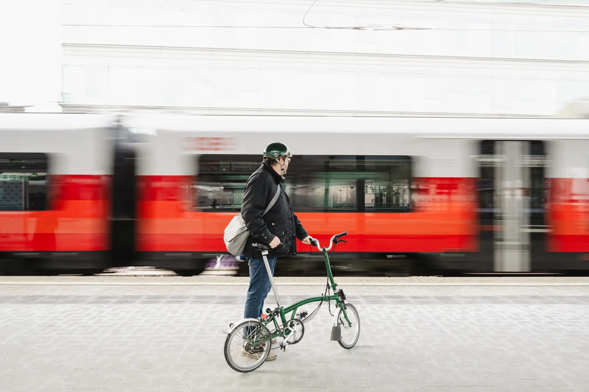 Person mit Faltrad steht am Bahnhof beim Gleis vor einem vorbeifahrenden Zug