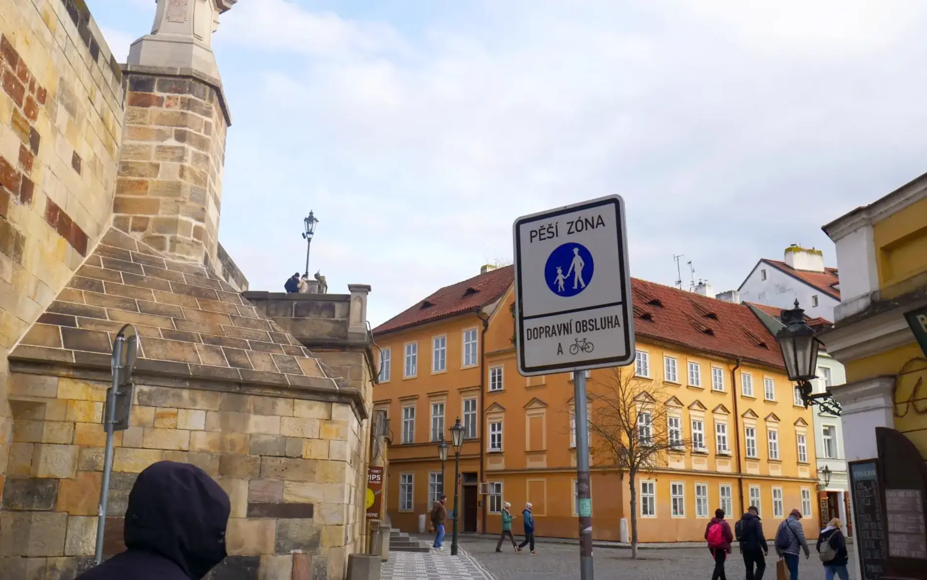 A cobblestone street in a historic area with beige buildings and a traffic sign in Czech indicating a pedestrian zone. Several people are walking near a stone structure on the left side of the image.