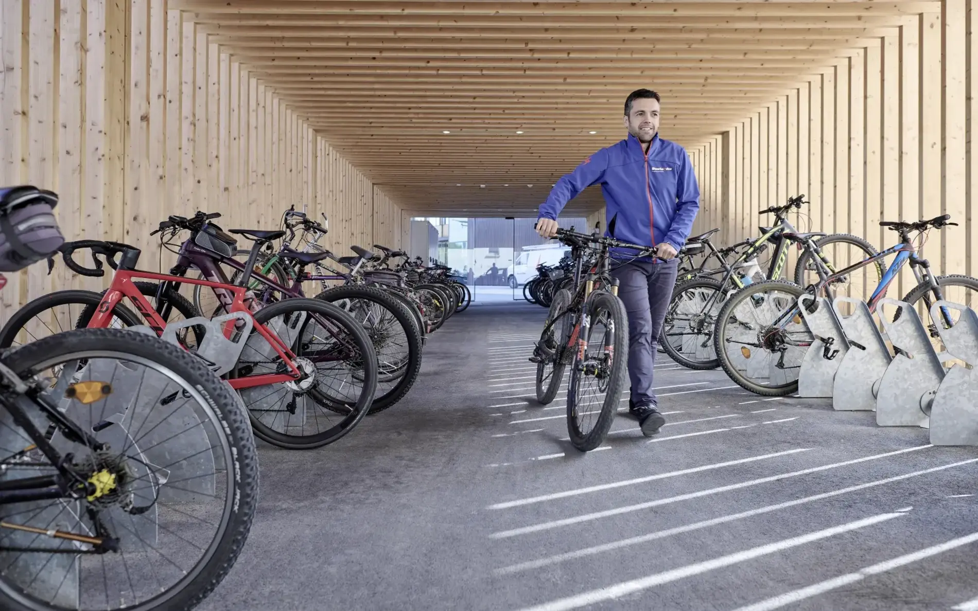 Ein Mann schiebt sein Rad in einen großen überdachten Radraum aus Holz. Links und rechts sind Räder in Rabstellanlagen abgestellt.