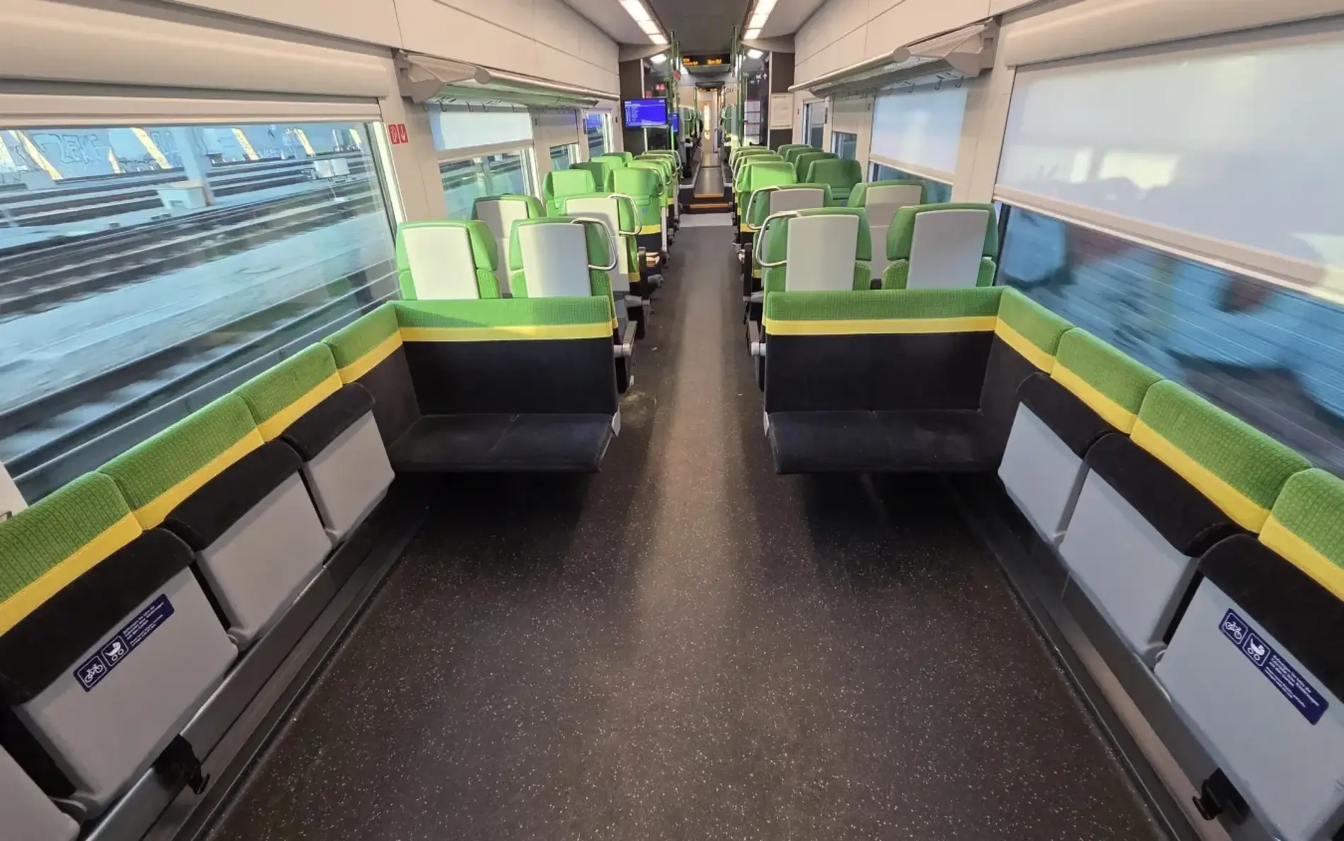 The interior of a modern train carriage with rows of green and gray seats arranged in pairs on both sides of a central aisle. Large windows line the walls, showing blurred tracks outside, and the space is brightly lit with overhead lights.