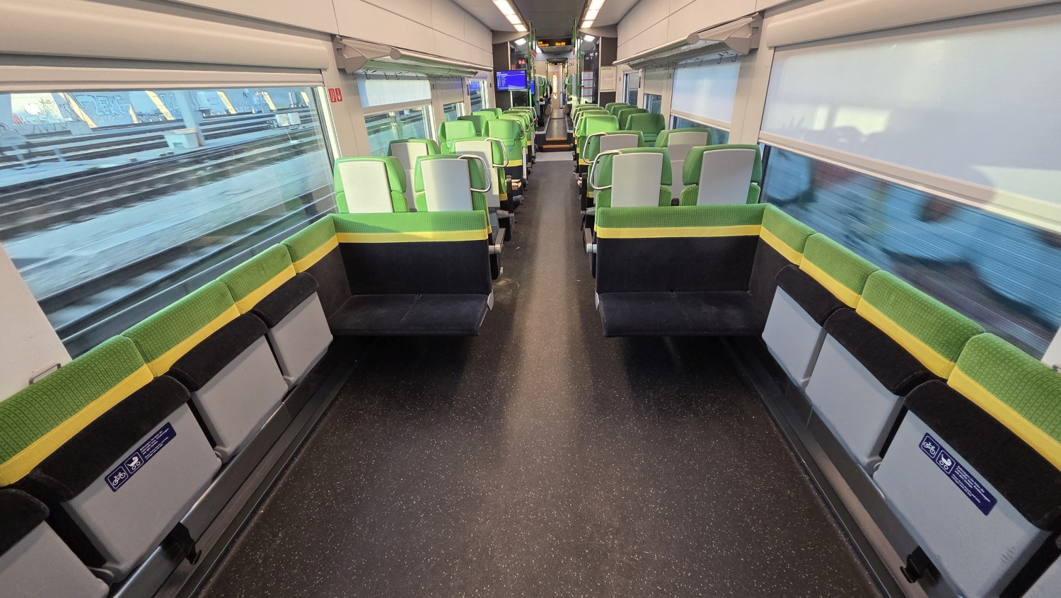 The interior of a modern train carriage with rows of green and gray seats arranged in pairs on both sides of a central aisle. Large windows line the walls, showing blurred tracks outside, and the space is brightly lit with overhead lights.