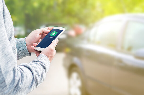 Hand hält Handy und eine App ist darauf zu sehen. Im Hintergrund ein Auto