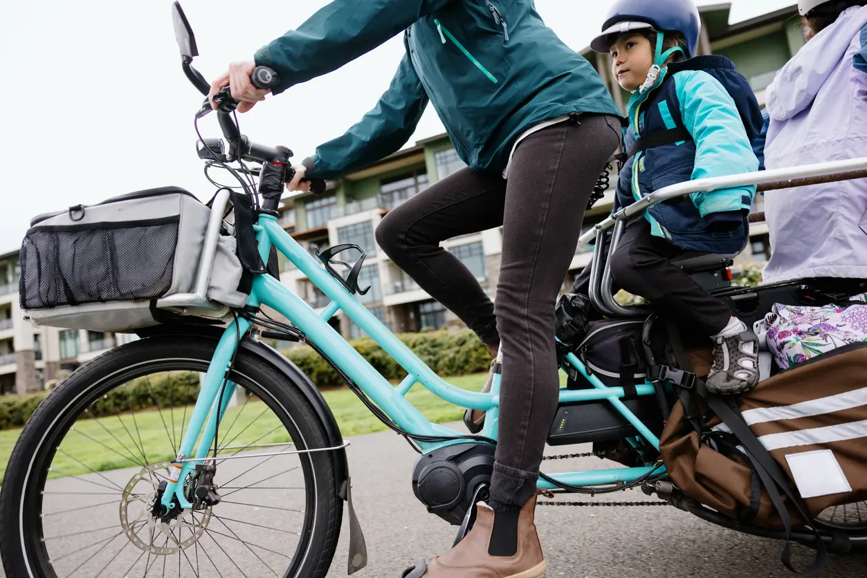 Zu sehen ist eine Frau, die ein Lastenrad fährt. Auf der Ladefläche sitzen zwei Kinder