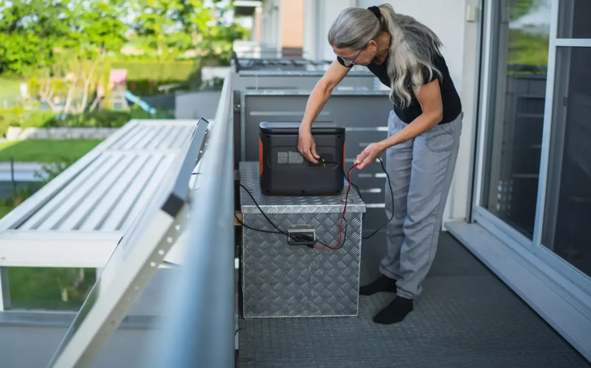 Eine Frau steht auf einem Balkon und schaut ihren Stromspeicher genauer an. Daneben ist ein Balkonkraftwerk.