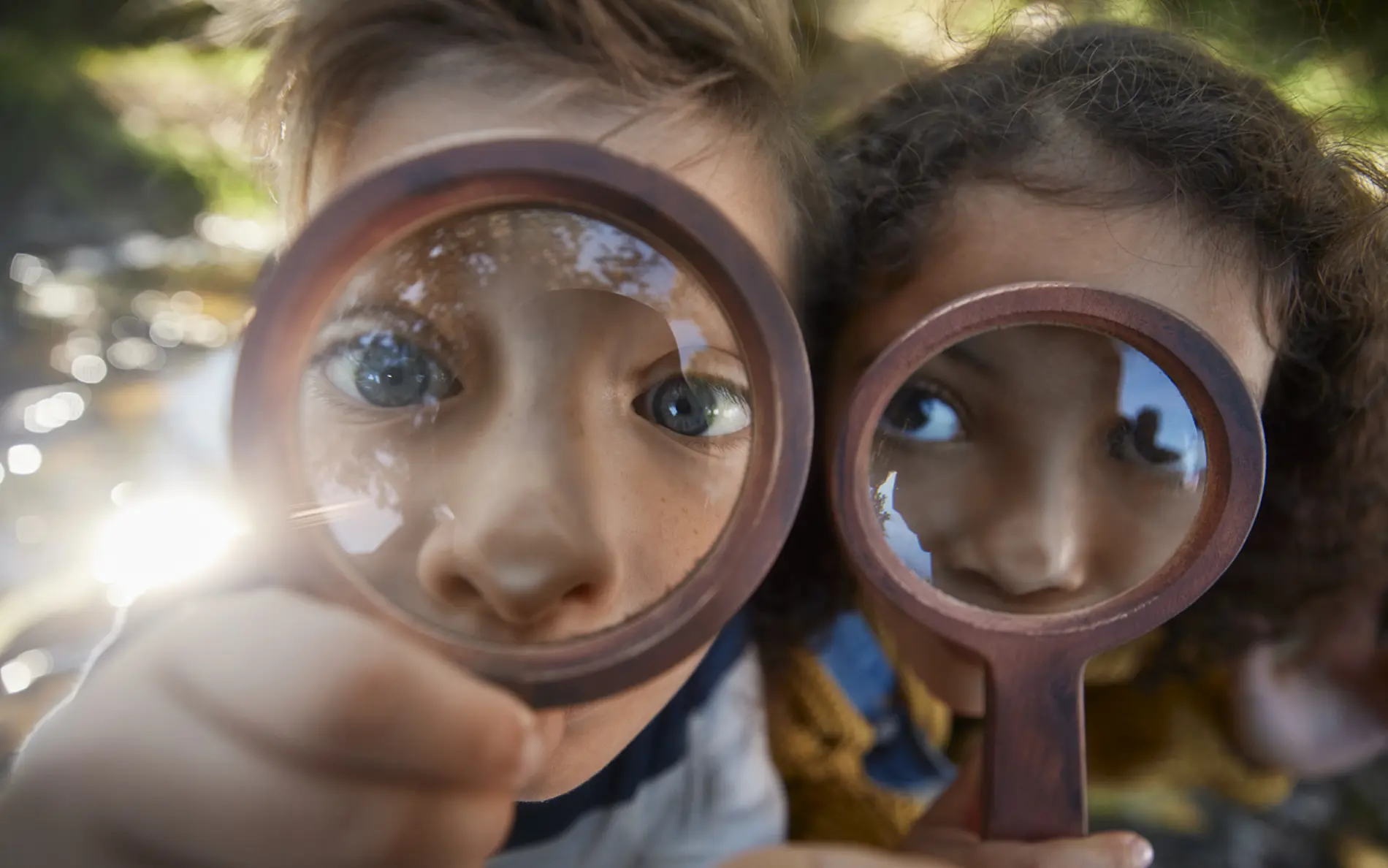 Nahaufnahme von zwei Kindern im Freien, die jeweils eine Lupe vor das Gesicht halten. Durch die Vergrößerungsgläser erscheinen die Augen und Teile der Gesichter deutlich vergrößert. Das Kind links hält die Lupe direkt vor ein Auge und blickt in Richtung Kamera. Das Kind rechts hält ebenfalls eine Lupe vor das Gesicht und blickt seitlich.  Im Hintergrund sind unscharfe natürliche Elemente zu erkennen, darunter Wasser, Pflanzen und Sonnenlicht. Die Aufnahme entstand bei Tageslicht und nutzt eine geringe Tiefenschärfe, wodurch die Lupen und Gesichter im Vordergrund scharf hervorgehoben werden.  Die Szene vermittelt das Thema Entdecken, Beobachten und Forschen in der Natur.