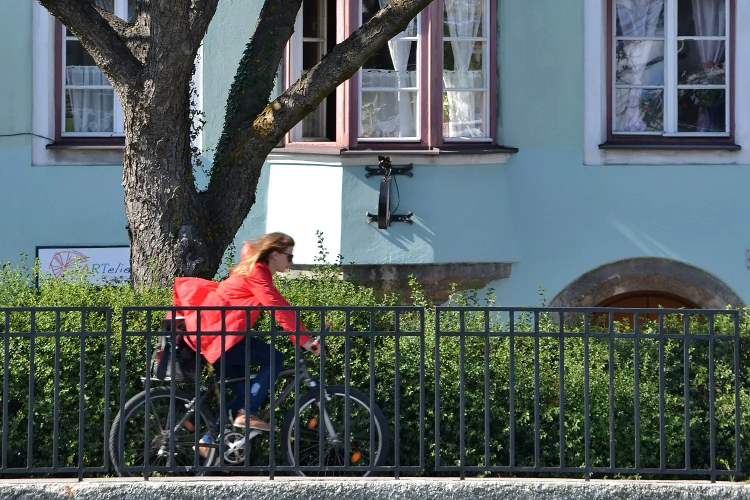 Radfahrerin vor Büschen, Bäumen und einem blauen Haus am radeln