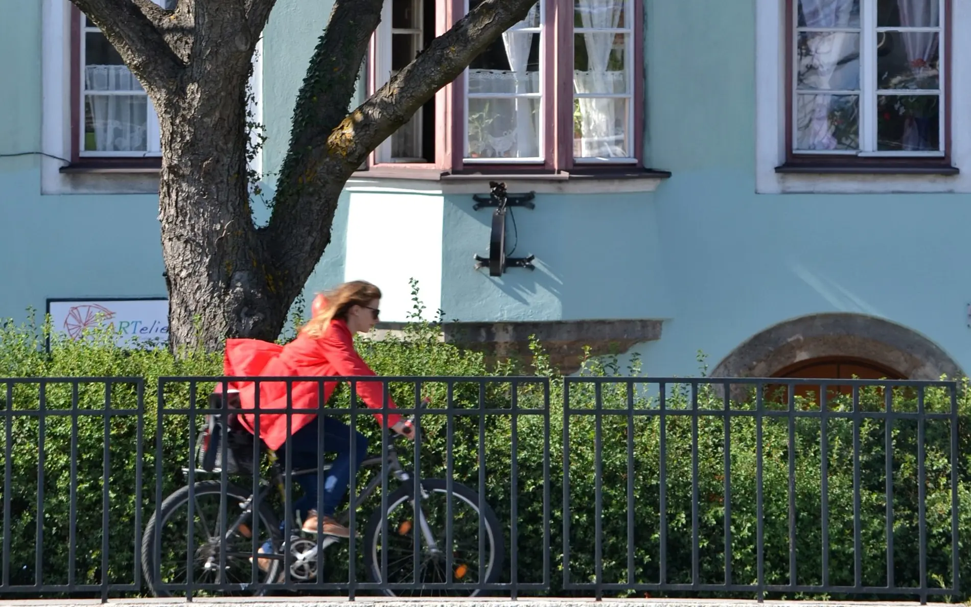 Radfahrerin vor Büschen, Bäumen und einem blauen Haus am radeln