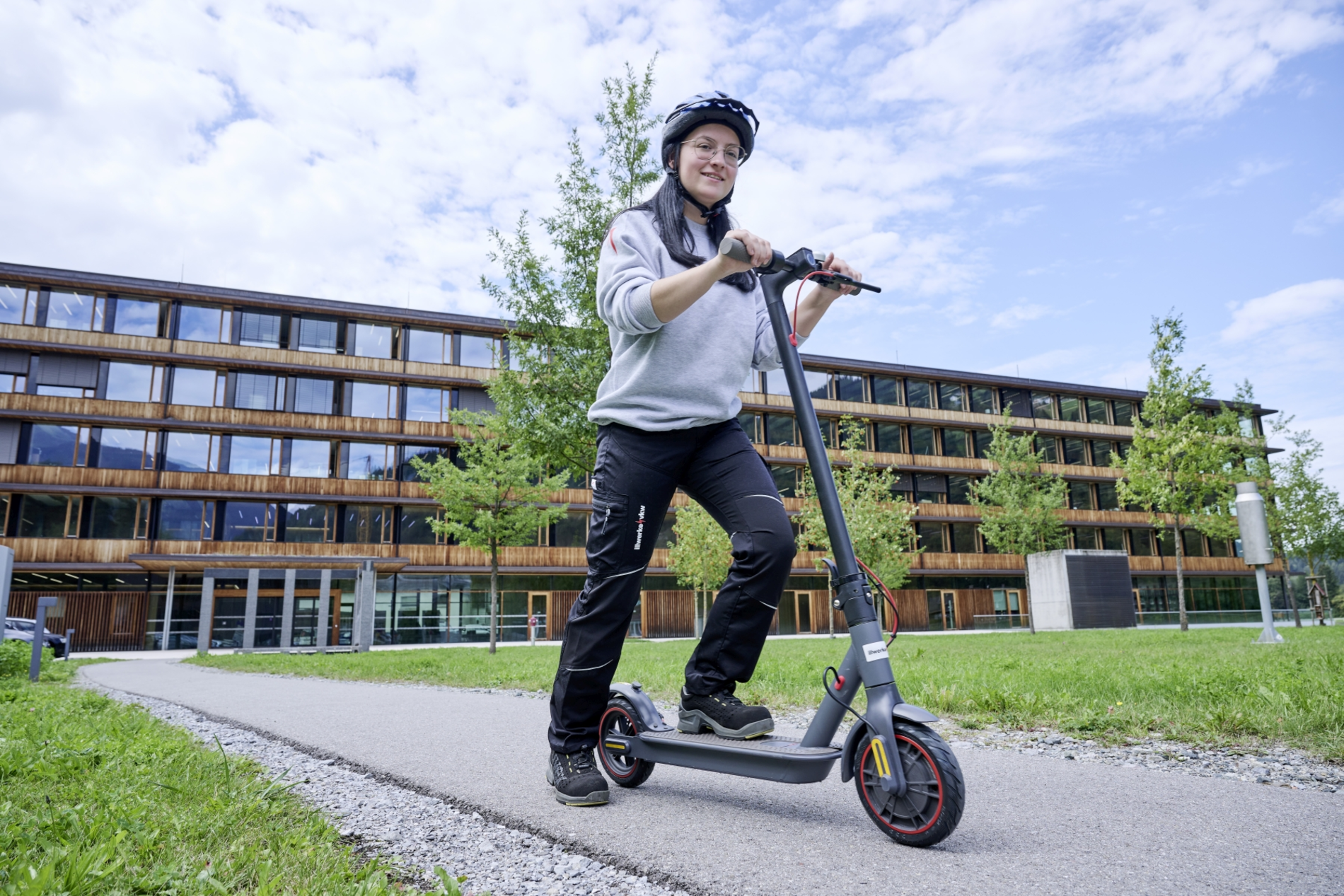 Frau mit Helm fährt Roller entlang eines Weges vor den illwerken. Das Gebäude im Hintergurnd ist aus Holz und Glas. Neben dem Weg reichlich Grünflächen