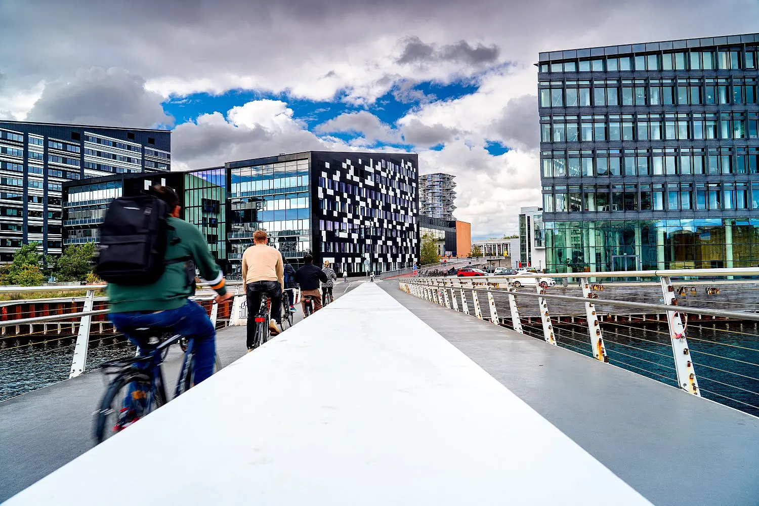 Radfahrer:innen über eine moderne Brücke in Kopenhagen, im Hintergrund moderne Glasgebäude