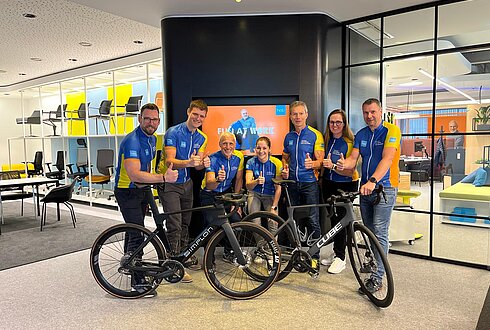 7 Menschen mit Fahrrädern, Innenraum eines Unternehmens, sie tragen Radbekleidung und zeigen "Daumen hoch"