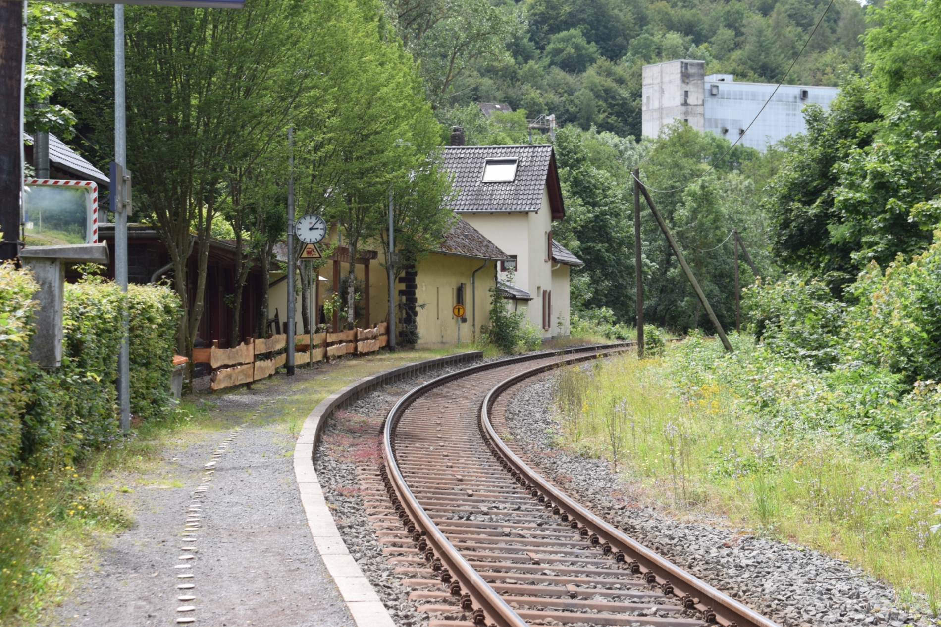 Ein älterer kleiner Bahnhof in der Ferne, vorne der Bahnsteig in einer Kurve. Die Gleise daneben schauen älter aus. Seitlich und im Hintergrund viel Grün