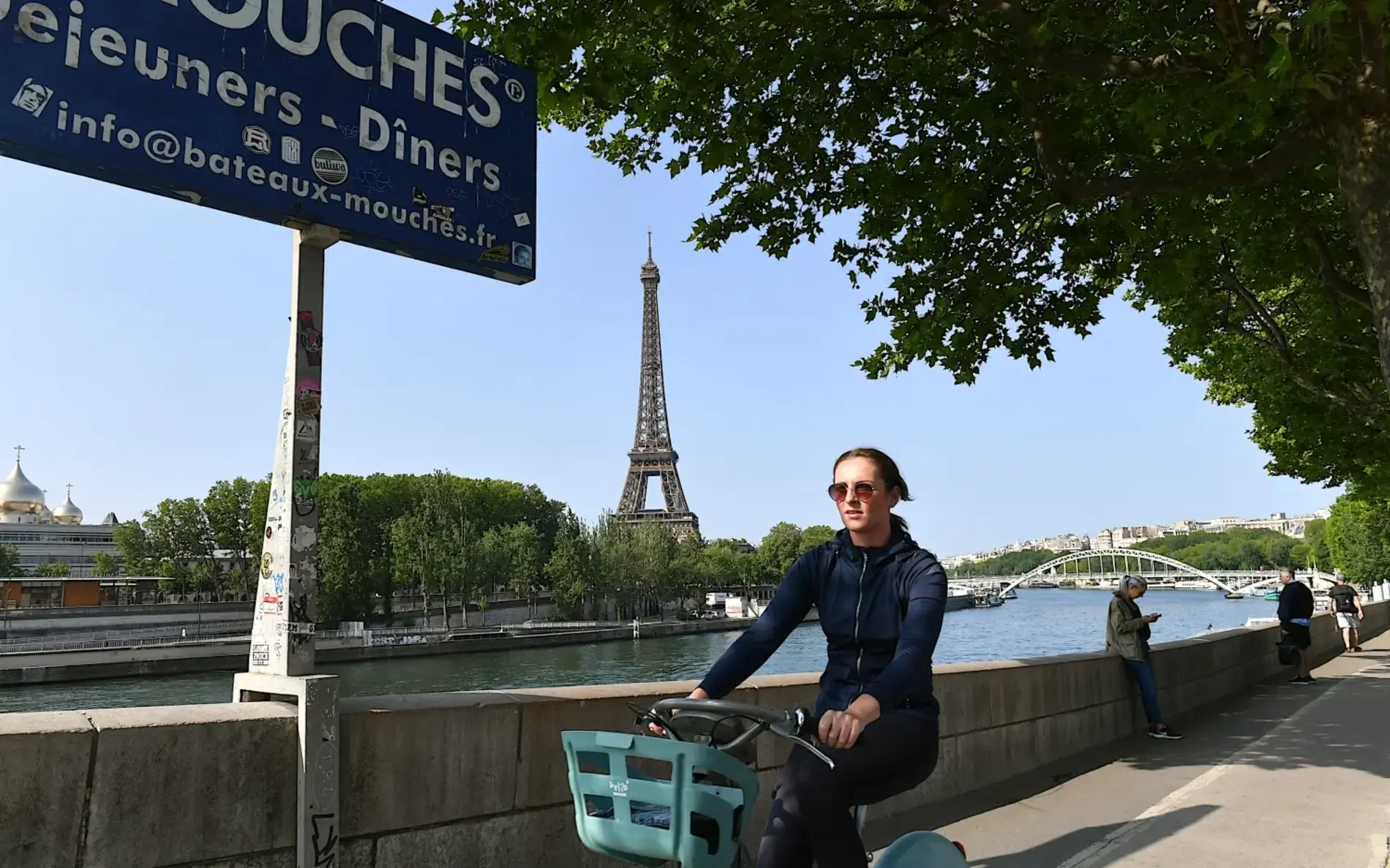 Junge Frau mit Sonnenbrille radelt entlang der Seine, im Hintergrund der Eiffelturm