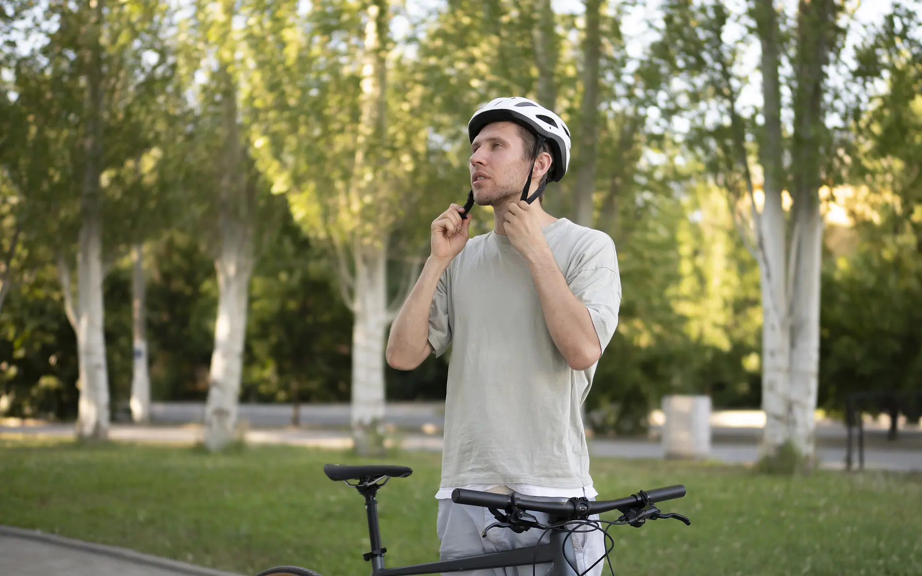 Mann setzt sich Fahrradhelm auf, sein Fahrrad angelehnt an seinen Körper. Im Hintergrund ein Park