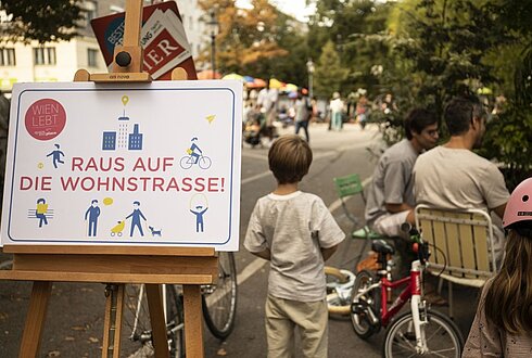 Schild auf Staffelei "Willkommen in der Wohnstraße". Rechts daneben steht ein Kind, ein Laufrad und mehrere Personen sitzend in Stühlen. Im Hintergrund sieht man vage eine Straße mit Bäumen und Spielsachen