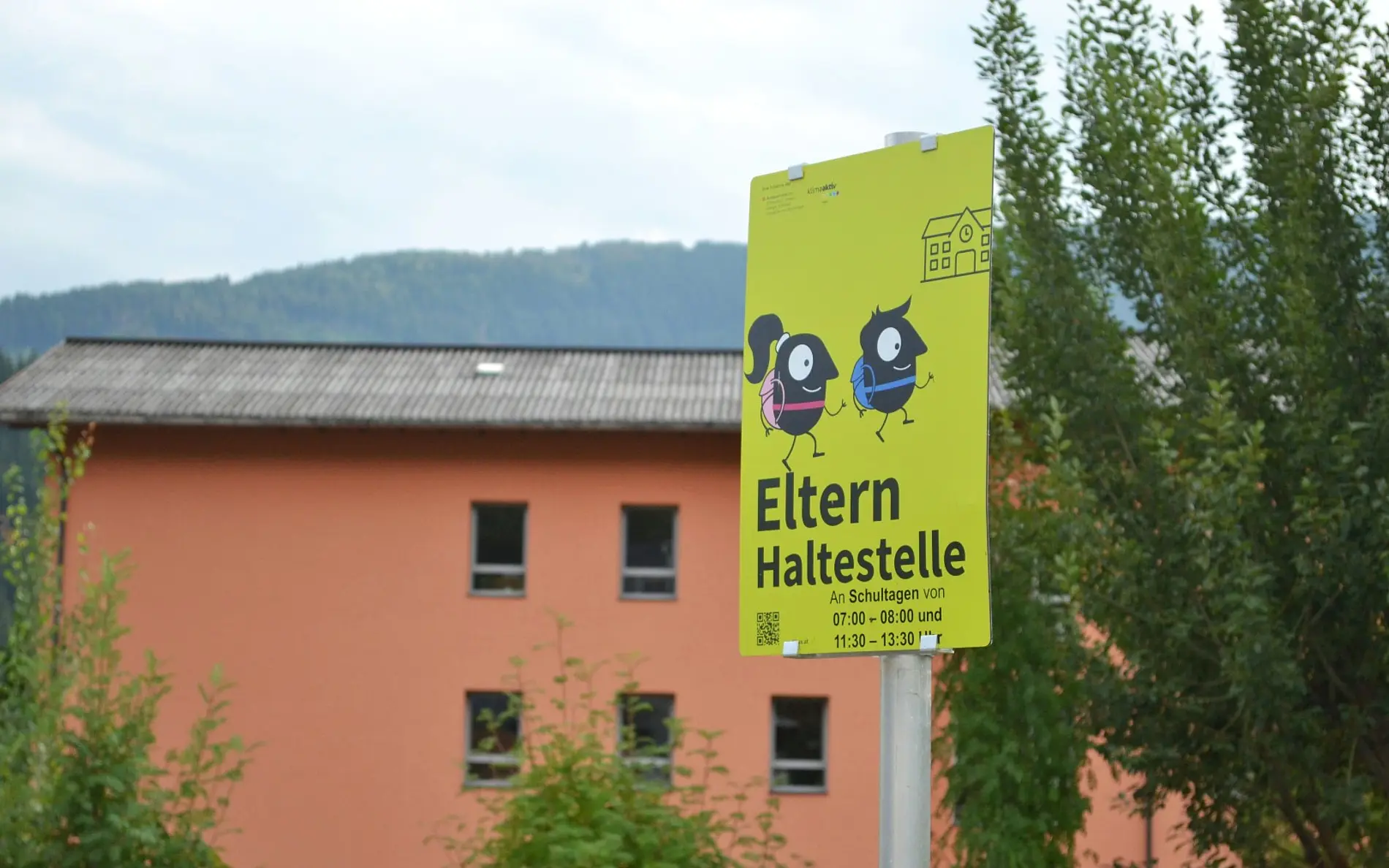 Schild Elternhaltestelle, im Hintergrund ein breites oranges Schulgebäude