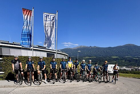 Viele Radfahrer:innen stehen nebeneinander mit gleichem Fahrradtrikot auf der Straße, dahinter wehen zwei Fahnen im Wind