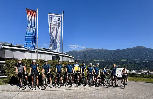Viele Radfahrer:innen stehen nebeneinander mit gleichem Fahrradtrikot auf der Straße, dahinter wehen zwei Fahnen im Wind
