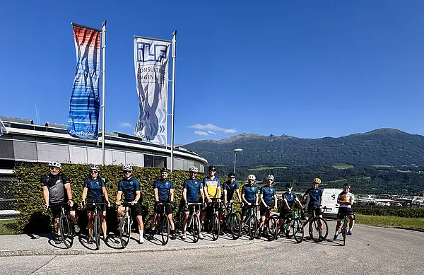 Viele Radfahrer:innen stehen nebeneinander mit gleichem Fahrradtrikot auf der Straße, dahinter wehen zwei Fahnen im Wind