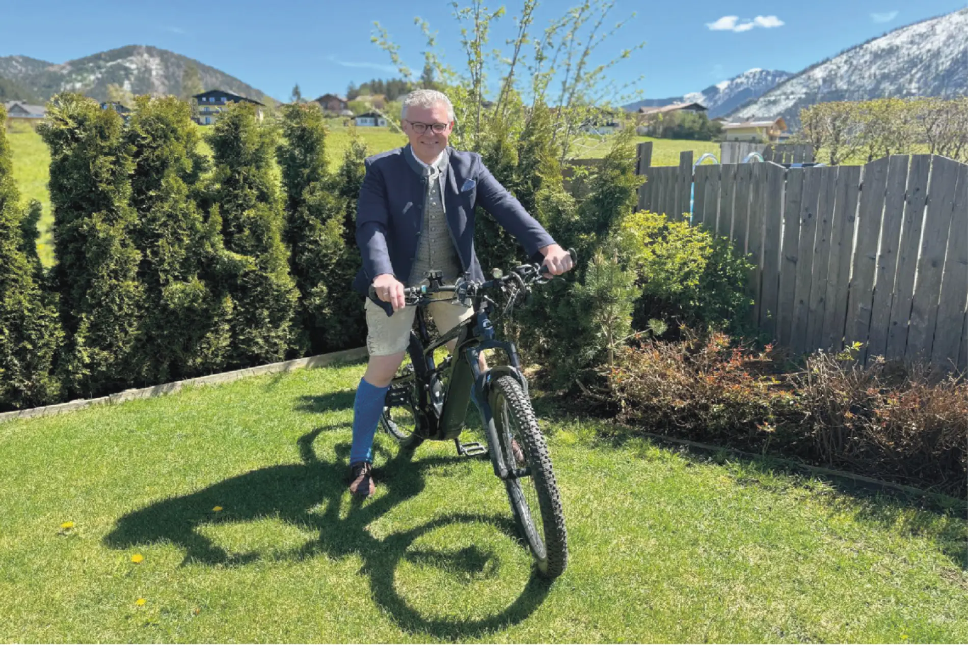 Wolfgang Ainz auf einem Fahrrad in einem grünen Garten. Im Hintergrund eine breite Berglandschaft.